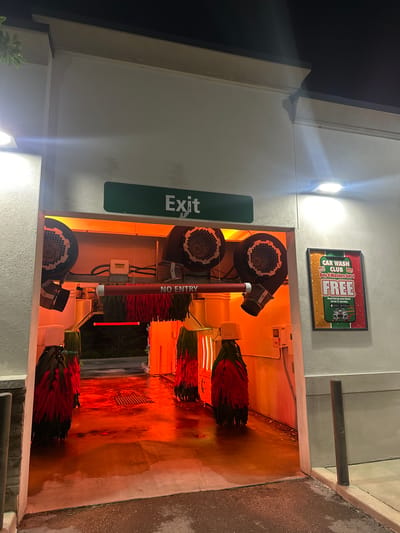 The image shows a car wash facility in Homosassa Springs, United States of America. The car wash has automated brushes and blowers for cleaning and drying vehicles. There is a sign that reads "Exit" next to a green rectangle. Another sign says "No Entry" near the equipment. A framed sign advertising the "Car Wash Club" is visible on the wall. The lighting inside the car wash is orange. The floor of the car wash is wet, and the surrounding area appears to be pavement. The image was taken at night or in a low-light condition.
