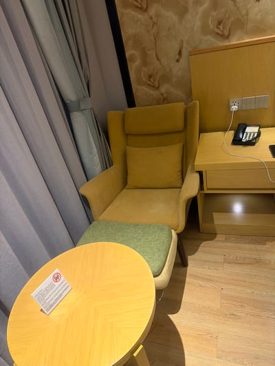 The image depicts a room interior. A yellow sofa with cushions is placed adjacent to a light wood veneer-covered cabinet structure. A green upholstered chair is positioned next to a circular wooden table, which bears a "no smoking" sign. The room's wall is paneled in light wood veneer on one side and a marble-like pattern on the other. Curtains hang down along the left side of the image. A black telephone is mounted on the cabinet. The location is not possible to be determined from the provided image.