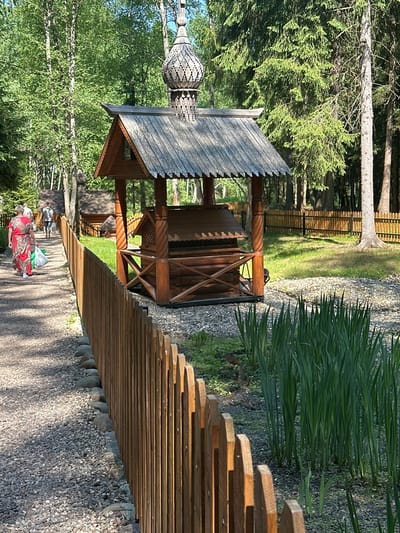 The image depicts a wooden gazebo with an ornamental top in Махра, Russia. A slatted wooden fence borders a pebble-covered path. Tall green grass grows alongside the path. In the background, there is a wooden fence and trees. A person wearing a red and white patterned garment walks along the path. Another person walks behind them. A second gazebo can be seen further back.