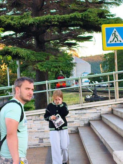 In Agoy, Russia, a man with short, dark hair and a beard, wearing a light blue t-shirt and a black backpack, looks to his left while standing on a concrete walkway. To his right and slightly behind him, a young girl with blonde hair, wearing a black jacket with white trim and grey sweatpants, holds a phone with a case depicting dogs, covering her face with her hand. Behind them, a pedestrian crossing sign is visible against a backdrop of lush green trees and a building. A train car can be seen in the distance. The scene appears to be outdoors, with a paved pathway and steps leading up to the walkway.