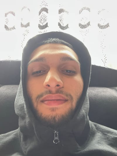 A person with dark hair and a beard is wearing a grey hooded sweatshirt. The person is facing the camera with their head slightly tilted. In the background, there is a white surface with repeating crescent shapes. The location is not discernible from the provided image.