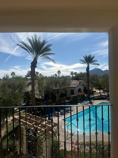 The scene depicts a sunny, clear day view from an elevated balcony in La Quinta, United States. The top and right edges of the frame show beige architectural structures, likely an overhang and a column. A black wrought iron railing with decorative scrollwork and vertical bars occupies the lower foreground. Immediately beyond the railing, a light blue swimming pool with curved edges is visible, its surface reflecting sunlight. Several blue lounge chairs and matching blue umbrellas are arranged on the light-colored paved deck surrounding the pool. A dark, low fence encloses the pool area. Behind the pool, a residential or resort complex features buildings with light stucco walls and red-tiled roofs, characteristic of Mediterranean or Spanish Colonial Revival architecture. Numerous palm trees, some very tall, and other green trees with dense foliage are interspersed throughout the complex. In the background, a range of mountains is visible under a bright blue sky, which contains scattered white clouds and prominent contrails forming an "X" pattern. No individuals are present in the image.