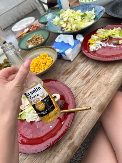 The image presents a top-down view of a casual dining scene, likely outdoors. A person's hands and lower arms are visible, one hand holding an empty "Coronita Extra" beer bottle. Several plates are arranged on what appears to be a wooden table. The plates contain various foods, including salads and corn. A box of tissues sits nearby. The presence of legs in the upper right suggests other people are present. The specific location in Jinhua, China, cannot be determined from the given visual information.