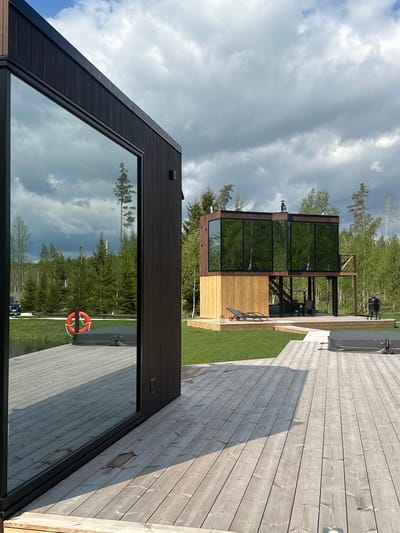 The image shows a contemporary architectural scene in Vaibla, Estonia. Two buildings are visible. The closer building has a dark, angular wooden exterior with a large window reflecting trees and the sky. A life preserver hangs on the adjacent wooden deck. Further back, a two-story building with large windows and a wooden frame stands amidst trees and manicured grass. A wooden deck connects the structures, featuring a hot tub and outdoor furniture. The sky is cloudy.
