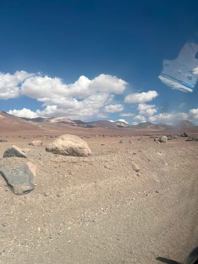 A bright blue sky is filled with puffy white clouds above a rugged desert landscape. The ground is covered in gravel and rocks, with larger boulders scattered across the terrain. In the distance, mountains rise against the horizon. A reflection of a window is visible in the sky, suggesting the picture was taken from inside. The horizon is on the left, with the desert extending right.