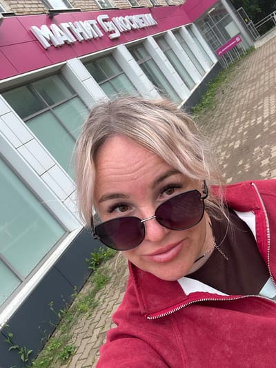 A woman is taking a selfie in front of a building with the sign "МАГНИТ КОСМЕТИК" in Chastyye, Russia. The building is red with large windows. The woman is wearing sunglasses and a red jacket. There is a bicycle parking sign visible. The ground is paved with cobblestones.