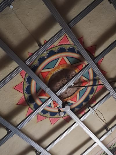 The image shows an overhead view of a ceiling with a circular, painted mural in the center. The mural features a sun-like design with radiating points in pink and yellow, concentric rings of color including blue, teal, and orange, and intricate dotted patterns. The ceiling is made of a light-colored material, possibly plasterboard, and is supported by a metal grid structure. A hole or dark recess is visible in the center of the mural. There are also electrical wires and what appear to be light fixtures attached to the metal grid. The context provided suggests this scene is located in Chobruchi, Moldova, though no specific elements in the image directly confirm this location. The overall impression is of an interior space, likely undergoing construction or renovation, with decorative elements on the ceiling.