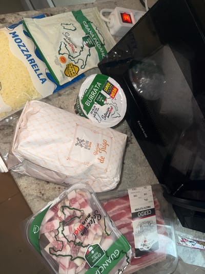The image depicts a collection of food items arranged on a speckled countertop, likely in a kitchen. The items include a bag of shredded mozzarella cheese, a package of Grana Padano cheese, a tub of burrata cheese, a bag of flour labeled "Harina de Trigo," and two packages of pork products, one labeled "GUANCIALE" and another with "CERDO" and "PANCETA FILETENDA". A black Taurus brand microwave is visible in the background, with a white power strip with a red illuminated switch. The lighting suggests an indoor setting. There are no people visible in the image. The scene appears to be staged in a domestic environment, possibly a kitchen counter in Sant Boi de Llobregat, Spain, given the presence of Spanish labeling on some products like "Selección Mundial" and "Origen Italia".