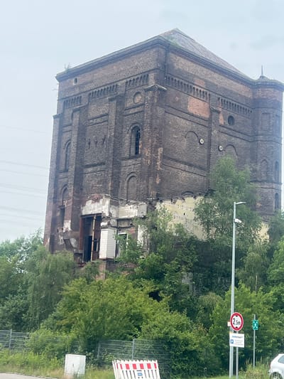 The image shows a tall, derelict brick building with a pyramidal roof. The building appears to be an old industrial structure, possibly a water tower, given its shape and the surrounding industrial landscape. The brickwork is dark and weathered, with sections of it crumbling and exposing the inner structure. There are several arched windows, some of which are broken or boarded up. The building is surrounded by dense green foliage, with trees growing up around its base and obscuring some of its lower sections. In the foreground, there is a metal fence, a white rectangular box, and a traffic barrier with red and white stripes. A lamppost stands near the fence, with a red circular sign showing "2.5m" and a green rectangular sign. The sky is overcast. The building is located in the industrial area of Herne, Germany.