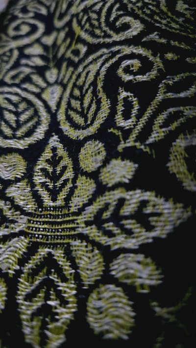 This is a close-up, high-angle shot of a black fabric with a yellow and white embroidered pattern. The pattern appears to be floral or paisley, with swirling shapes and leaf-like designs. The embroidery is textured and slightly raised, with visible threads. The fabric is likely a textile, possibly a rug or a decorative cloth, given the intricate design. The lighting is dim, creating shadows that emphasize the texture of the embroidery and the depth of the fabric. There is no visible text, people, or activity depicted in the image. The focus is entirely on the decorative surface of the textile.