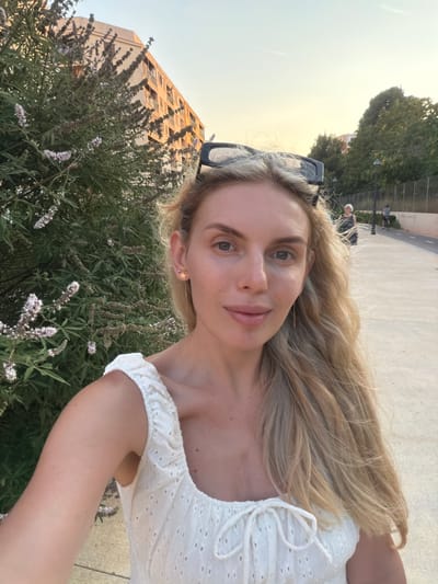 This is a portrait-oriented, full-body, outdoor selfie taken by a young Caucasian woman with long, blonde, wavy hair and light green eyes. She is wearing a white, off-the-shoulder top with eyelet detailing and a drawstring closure at the bust. A subtle star earring is visible in her left ear. She is smiling and looking directly at the camera, with a natural and pleasant expression.
Behind her, in the foreground, is lush green foliage with delicate purple flowers. Further back, to her left, a tall, multi-story building with visible windows and balconies is partially obscured by the greenery. To her right, a paved walkway can be seen, with several figures walking in the distance.
The lighting suggests it is late afternoon or early evening, with a warm, soft glow illuminating the scene. The sky is a pale yellow, indicating a sunset or sunrise. The overall atmosphere is relaxed and casual, possibly indicating a leisurely stroll or a moment captured during an outing. The location is identified as Benimàmet-Beniferri, Spain. No text is visible in the image.