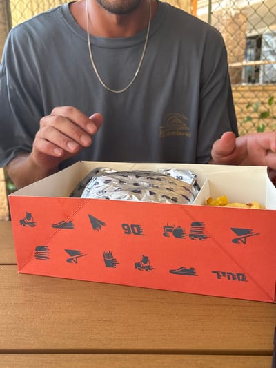 A close-up, slightly overhead view depicts a red and white paper food box resting on a light brown wooden table in an outdoor or semi-outdoor setting within the Galil Elyon Regional Council, Israel.
One individual, a male with visible stubble on the chin and neck, is partially visible behind the food box. He is wearing a charcoal grey short-sleeved t-shirt featuring a gold-colored "Quiksilver" logo on the left chest and a thin silver chain necklace. His hands are positioned near the top edge of the food box, fingers slightly curled.
The food box's red exterior is decorated with black silhouette icons including a roller skate, a running shoe, a paper airplane, the number "90", a tractor, a truck-like vehicle, a skateboard, a hamburger, a wheelbarrow, and french fries, each with implied motion lines. Hebrew text is also present. Inside, the box is divided into two compartments. The left compartment contains a food item, likely a burger, wrapped in silver foil with black graphics including the number "90". The right compartment holds yellow, irregularly shaped fried potato pieces.
The background is out of focus, revealing a light-colored wall or structure on the left, and a metal mesh or chain-link fence on the right, behind which are light-colored surfaces and green foliage. The scene is illuminated by natural light.