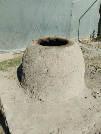 An outdoor scene in Yangibazar, Uzbekistan, shows a traditional tandoor oven made of clay. The oven is bell-shaped and sits on a concrete base. The surface of the tandoor is textured with visible cracks and embedded straw. Behind the tandoor, a light-colored tarp or fabric is stretched taut, casting shadows from an unseen object. The ground around the tandoor is composed of dirt and scattered dry grass. The lighting suggests it is daytime.
