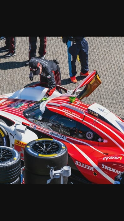In Oreshki, Russia, at an outdoor event, a race car driver in a black and red racing suit is bending over the side of a red and white race car. The driver wears a black cap and gloves. Two other individuals in similar racing attire stand nearby, one on the left and another on the right. In the foreground, stacked racing tires are visible, and a small green object is placed on the car's roof. The ground appears to be paved.