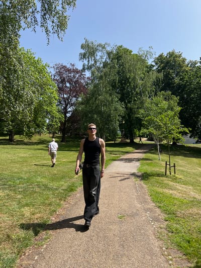 The image depicts a park scene, likely in Ipswich, United Kingdom, given the location provided. A light-skinned male is walking on a gravel path in the foreground; he wears a black tank top, dark pants, and sunglasses. He appears to be holding a dark-colored object, possibly a cell phone, in his right hand. His shadow extends onto the path and the adjacent grass. In the background, a second light-skinned individual, wearing a light-colored shirt and shorts, walks on the lawn parallel to the path. The environment consists of green grass, mature trees, and other shrubbery, suggesting a park or recreational area. A clear blue sky is visible through the branches of the trees. A small, newly planted tree is held in place with wooden stakes.