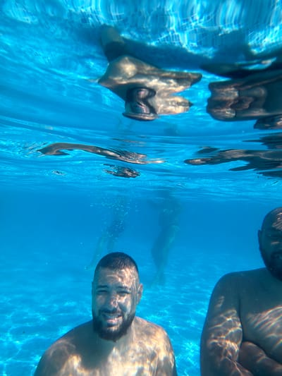 An underwater photograph shows three people in a swimming pool. The person in the foreground on the left is a man with a beard and short hair, smiling at the camera. To the right of him, another person's head and shoulder are visible, also looking towards the camera. In the background, a third person is swimming away. The water is clear blue, and sunlight creates patterns on the bottom of the pool. The image appears to have been taken in the city of Chiflik, Bulgaria.