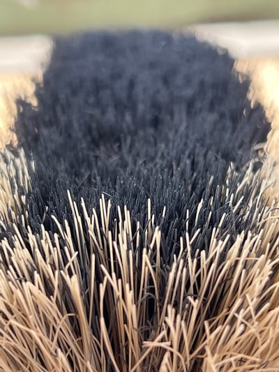 This is an extreme close-up, macro photograph of the bristles of a brush, likely a hairbrush or a cleaning brush, taken outdoors. The bristles are arranged in dense tufts, with a mix of dark, almost black bristles and lighter, straw-colored bristles. The focus is sharp on the tips of the bristles, revealing their texture and individual strands. The background is blurred and out of focus, showing a soft green hue at the top, suggesting a natural, possibly grassy environment. The scene is static, with no people or actions depicted. The lighting appears to be natural daylight, creating subtle highlights and shadows on the bristles. The photograph does not contain any information that would suggest a location within the city of Lanžhot, Czechia.
