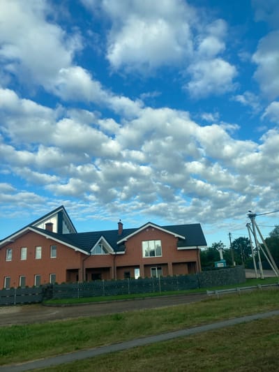 A bright, wide shot captures a modern brick house with a dark roof under a cloudy blue sky. The house has multiple windows, some reflecting the sky. A dark fence encloses the property, with green bushes visible behind it. To the right, there are utility poles and trees. In the foreground, there is a grassy area with a paved path. The scene is located in the Матыкальскі сельскі Савет, Belarus.