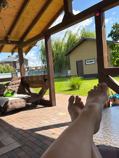 A person's bare feet and lower legs are positioned in the foreground, crossed at the ankles, resting on a glass-topped table. The perspective is from the person's point of view, looking out from a covered patio or gazebo. The patio has a wooden roof with exposed beams and wooden posts. Below the roof, on the left, a brown cushioned sofa is visible with a white throw blanket and a bright green electric tool. The patio floor is made of brick pavers. In the background, a well-maintained green lawn is visible, with a small building that appears to be a garage or shed, painted a light olive color. A large weeping willow tree with green foliage dominates the middle ground. Beyond the lawn, houses with pitched roofs and a fence can be seen. The sky is blue with scattered white clouds. This scene is located in the village of Shopino, Russia.