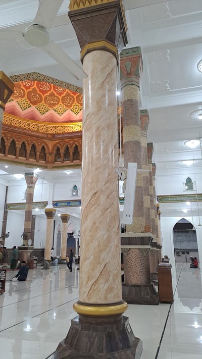 The image shows the interior of a mosque in Lhokseumawe, Indonesia. The foreground is dominated by a large, ornate pillar with a marble-like finish. Behind it, several other pillars of varying designs, some with mosaic patterns, are visible. The ceiling features intricate decorative patterns, including Arabic calligraphy in gold and red. Several ceiling fans are also visible. The floor is a highly reflective white, with dark lines indicating different sections. People are scattered throughout the mosque, some sitting on the floor, others walking. Some individuals appear to be praying. The overall impression is one of a spacious and well-decorated place of worship.