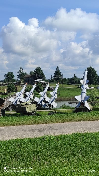 The image shows a collection of military missiles on display outdoors. In the foreground, there are five missiles, four of which are arranged in a row on a mobile launcher. The fifth missile is positioned to the right, also on a launcher. The missiles are white with orange tips and have distinct fin structures. Behind the missiles, there is a body of water, likely a pond or lake, with a grassy bank. Further in the background, there are trees, buildings that appear to be part of a museum or exhibition, and more military vehicles, including what looks like a tank. The sky is blue with scattered white clouds. The ground is covered with green grass, and a paved path runs through the scene. The date and time stamp on the image indicate it was taken on July 15, 2026, at 10:29. The location is identified as the village of Лашанскі сельскі Савет, Belarus, suggesting this is likely an outdoor museum or memorial site. No people are visible in the image.