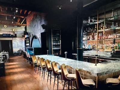 The media file shows an interior view of a dimly lit, upscale bar or restaurant. The scene is empty of people. The focus is on the bar area, which features a long, polished stone countertop supported by a textured base. Several bar stools with plush, golden upholstery and dark wood legs are neatly arranged along the counter. Behind the bar, shelves are filled with numerous bottles of liquor and glassware, illuminated by subtle lighting, suggesting an evening setting.
To the left of the bar, tables and chairs are visible in the foreground, also empty. Further back, an elevated mezzanine level with railings can be seen, adding depth to the space. The walls have a textured, mural-like design in muted tones, and the ceiling reveals exposed wooden beams and industrial-style lighting fixtures. The overall ambiance is sophisticated and modern, with a hint of industrial chic. There is no visible text in the image. The location context provided is Ellwangen, Germany, but there are no specific visual cues within the image to confirm this. The time of day appears to be evening or night, given the artificial lighting and the absence of natural light.