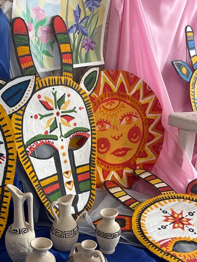 The scene depicts an indoor display of folk art elements, located in Tayshet, Russia. The primary subjects include multiple brightly painted, stylized cutouts and a collection of ceramic pottery.
In the central foreground, a large, white, mask-like cutout with black-outlined eye holes, a small mouth slit, and geometric patterns in red, green, yellow, and black is prominently displayed. It is adorned with two upward-curving, striped ear-like appendages. To its immediate right, a circular cutout portrays a stylized yellow sun with a human-like face rendered in red and orange tones, surrounded by radiating sunbeams. Partial views of additional painted cutouts are visible to the left and lower right, including one circular piece featuring a red and yellow starburst design, along with striped appendages similar to the main mask. These cutouts appear to be made from a rigid material, possibly cardboard.
Arranged on a blue fabric in the lower foreground are approximately six unglazed, light-colored ceramic vessels of varying sizes. These include pitchers with handles and smaller open-mouthed vases, many adorned with a dark, maze-like or geometric pattern around their midsections.
The background consists of a blue fabric drape on the left, two distinct watercolor paintings (one depicting pink roses, the other purple irises) centrally placed, and a draped pink satin fabric on the right. A partial view of a light-colored, possibly wooden, stool or small table is visible under the pink fabric on the far right. A translucent white netting or sheer fabric is draped throughout the display, particularly over parts of the background and foreground. The items are arranged in a static configuration, indicative of an exhibition or decorative setup.