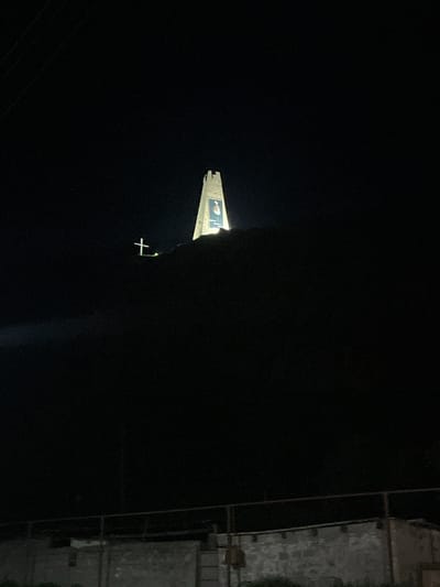 The image depicts a nighttime scene in Sioni, Georgia, featuring a prominent tower illuminated against the dark sky. The tower is topped with crenellations, and a large portrait is displayed on its side. To the left of the tower, a illuminated cross stands on a dark hillside. In the foreground, a low, stone wall with a metal railing runs horizontally across the frame, suggesting a boundary or a viewing area. The overall environment is dark and appears to be an open, possibly elevated area.
