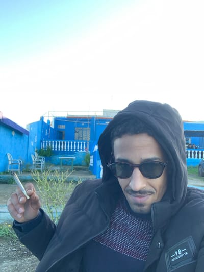 A man in a black hooded jacket and sunglasses holds a cigarette in his hand. He is positioned in front of a bright blue building with white accents, including a railing and stacked chairs. The sky is overcast, and the ground appears to be paved. The man is facing the camera, with a slight smile. A patch on his jacket reads "-18-".