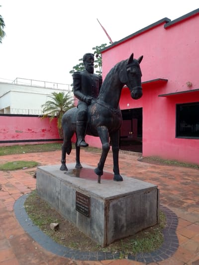 This image presents an equestrian statue of General Bernardino Caballero, located in Concepción, Paraguay. The dark metal statue depicts a man with a prominent beard and mustache, dressed in a formal military uniform with epaulets, riding a horse. The horse is shown in a dynamic pose, with one front leg slightly raised.

The statue is mounted on a rectangular, grey concrete pedestal. A bronze plaque on the front of the pedestal provides text details: "GRAL. BERNARDINO CABALLERO / OBRA DONADA POR EL / DR. BERNARDO VILLALBA CARRIZO / AL DR. OTILIO ROLAND GONZALEZ / 11 ABRIL 2011". This identifies the subject as General Bernardino Caballero and details the donation and dedication on April 11, 2011.

The setting includes a ground covered with reddish-brown pavers, with sections of grass and dirt visible around the statue's base and between the pavers. In the background, to the left, there is a white building with a flat roof, partially obscured by palm trees and a fence topped with barbed wire. To the right, a vibrant pink or magenta building with a flat roof and dark-framed windows provides a striking contrast. Black trim is visible along the roofline and under some windows of this building, and a doorway is open on its right side.

The scene is illuminated by natural daylight, suggesting an overcast or brightly diffused sky, as there are no strong shadows. The overall impression is one of historical reverence within a somewhat rustic outdoor public space.