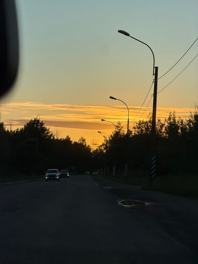 The image depicts a road at sunset in the city of Pontonny, Russia. The sky is a gradient of light blue and warm orange hues, with distinct horizontal cloud formations catching the setting sun's light. Silhouetted against the sky are trees lining both sides of the road, creating a dark, dense border. Several cars are visible driving on the asphalt road, with their headlights illuminated, suggesting the approaching twilight. Streetlights, also silhouetted, are spaced along the right side of the road, some with curved arms. The road surface shows signs of wear, including a few puddles reflecting the sky's colors. The overall impression is of a tranquil, late evening drive on a rural or semi-urban road.