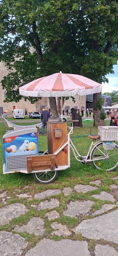 An outdoor scene in Kuressaare, Estonia, captures a mobile ice cream cart parked on a grassy area with stone paving in the foreground. The cart, a white bicycle modified with a wooden structure and an ice cream freezer, is adorned with a pink and white striped umbrella. Behind the cart, a man in a dark jacket and jeans stands looking towards the right. In the background, a large stone building, possibly a castle, is visible. Several vehicles, including white vans and cars, are parked in front of the building. A crowd of people is gathered in the distance, suggesting an event or gathering. The overall atmosphere appears to be a pleasant, possibly evening, outdoor setting.