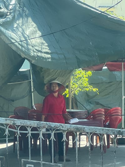 A person in a conical hat and red clothing sits amidst a collection of stacked red plastic chairs near a window with a white metal frame. A large gray tarp covers part of the scene, obscuring the background. Hints of green foliage are visible behind the tarp and chairs. The image is slightly tilted, giving a distorted perspective of the scene.