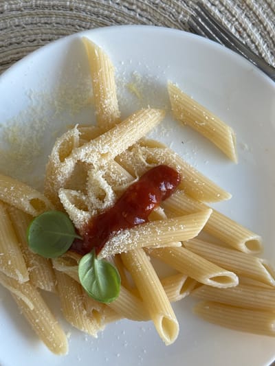 A close-up, overhead shot captures a white plate filled with cooked penne pasta. The pasta is sprinkled with grated cheese and adorned with a dollop of red sauce and two fresh green basil leaves. The plate rests on a textured, light brown placemat, and the corner of a silver fork is visible in the upper right of the frame. The scene is lit from above, casting subtle shadows. The image does not provide any information about the location in the city of Гамово, Russia.