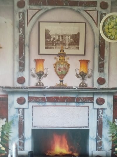 This is a photograph of a decorative fireplace mantel with a large framed picture of the White House above it. The mantel is adorned with two ornate lamps with orange glass shades, and a decorative vase. To the right of the picture is a round clock with what appears to be fruit for numbers. The fireplace itself is visible below the mantel, with a fire burning within. The overall style suggests an indoor setting, possibly a living room or dining room. The prompt mentions a specific location in Belarus, but there are no visual cues in the image to support or refute this.