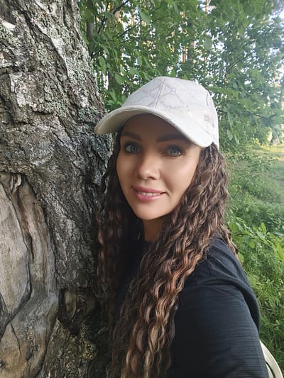 A woman with long, curly brown hair and blue eyes is pictured from the chest up, leaning against a large tree. She is wearing a white baseball cap with a patterned design and a black shirt. The background consists of green foliage and trees, suggesting an outdoor, natural setting. The image appears to be a selfie taken outdoors, possibly in a park or wooded area. There is no indication of the specific location within the city of Chaikovsky, Russia.