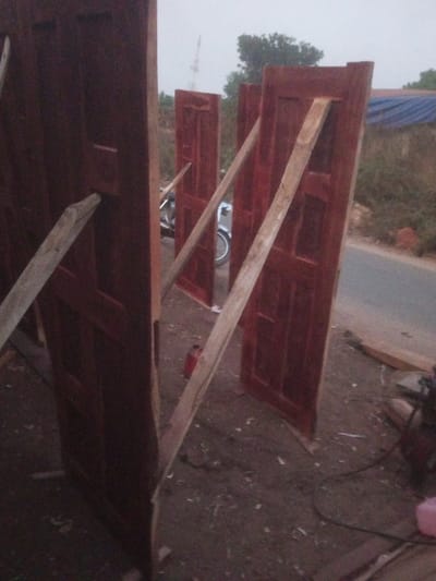 This image captures an outdoor scene in Ebiya, Nigeria, depicting several newly finished or processed wooden panel doors standing upright. There are at least four reddish-brown doors, each supported by light-colored wooden planks angled as braces. The foreground ground is composed of dark, disturbed earth mixed with wood shavings and debris. In the mid-ground, a paved road or pathway is visible to the right. A motorcycle or bicycle is partially discernible behind one of the central doors, near the road. Further in the background, sparse green vegetation and a tall, thin antenna-like structure are visible. A blue tarp-covered object is also present behind the doors on the right side. On the ground, a red cylindrical container and other indeterminate items, including a pink container, are present. No individuals are visible in the scene. The lighting suggests overcast conditions or low light.