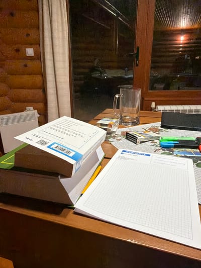 The scene depicts a table indoors, situated near a window. A clear glass mug is present, along with various items including cigarettes, markers, a notepad, and what appears to be a stack of documents or paperwork. The location is identified as Stoikite, Bulgaria, based on information visible on one of the documents. The table is wooden, and the background shows elements of a room with wood paneling. The scene appears still, with no visible people or actions taking place. The environment is a room with a mix of personal and professional items.