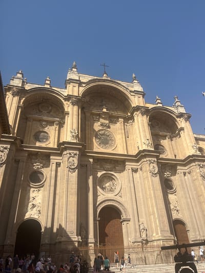 The image shows the exterior of the Granada Cathedral in Granada, Spain. The perspective is from a low angle, looking up at the ornate facade of the cathedral against a clear blue sky. The cathedral's architecture is characterized by its large arched windows, detailed sculptures, and classical design. Sunlight casts strong shadows across the stonework, highlighting the architectural features.
In the foreground, a crowd of people is gathered on the steps leading up to the cathedral. They appear to be tourists, with many holding cameras or looking towards the building. Some are walking, while others are standing and talking. A metal fence separates the gathered crowd from the cathedral entrance. The overall environment suggests a sunny day in a popular tourist location.