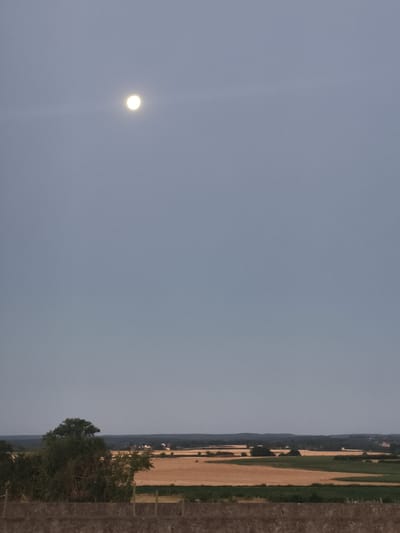 The image captures a twilight landscape, dominated by a pale, hazy sky with a prominent full moon positioned in the upper left quadrant. Below the sky, a rural panorama unfolds, featuring cultivated fields with dry, golden stubble and patches of green vegetation. A line of trees, some dense and dark, others sparser, forms a middle ground, with a few small buildings visible amongst them. In the foreground, a low, textured stone wall runs horizontally across the bottom of the frame. The scene appears to be an overlook of the countryside, potentially near the village of Époisses, France, given the agricultural setting and distinct rural architecture.