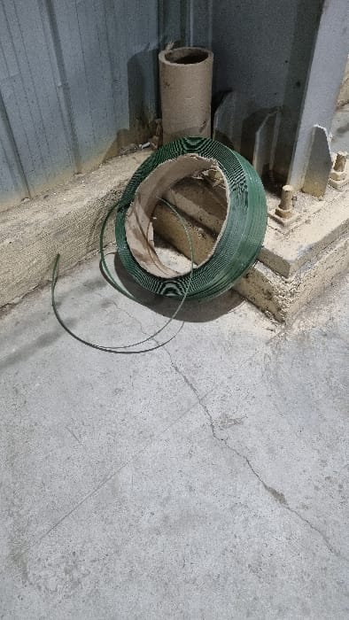 The image displays a close-up view of industrial or discarded materials within what appears to be an indoor or covered outdoor setting in Chapyk, Uzbekistan. The central subject is a partially used coil of green strapping material, likely made of plastic, resting on a textured surface. A length of this green material extends loosely from the coil. Adjacent to the coil, a brown, cylindrical cardboard tube is visible, positioned horizontally.
The surroundings consist predominantly of aged, grey concrete, exhibiting several visible cracks that suggest wear or settlement. To the left, a section of corrugated metal is present, possibly a wall panel or part of a roof structure. Above this, a robust, grey metal framework or beam is partially visible. The lighting indicates it is daytime. There are no people, explicit activities, or visible text in the frame. The scene conveys a sense of a storage area, a construction site, or a utilitarian space.