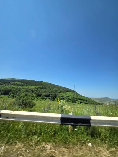 The image shows a hilly, green landscape under a clear blue sky. A white and black guardrail is in the foreground, with blurred greenery and dry grass beneath it. In the mid-ground, a lush green hillside is covered with trees and grass, with several tall, slender transmission towers visible on its slope. In the distance, rolling hills fade into the horizon. There are no people or animals visible in the image. The location is the city of Krashen, Armenia.