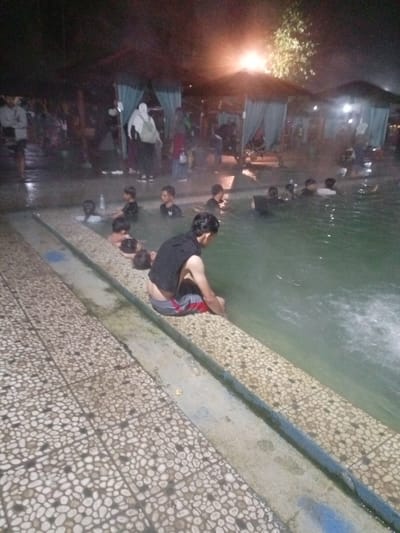 This nighttime image captures a lively scene at a swimming pool or hot spring in Kecamatan Pangalengan, Indonesia. Numerous individuals, predominantly men and boys, are either submerged in the water or gathered around its edges. The water appears to be warm, with visible steam rising, suggesting it may be a hot spring or heated pool.
In the foreground, a man sits on the tiled edge of the pool with his feet immersed in the water. He wears red and grey striped shorts and a dark sleeveless top, with the back of his shorts slightly pulled down. Other people are dispersed throughout the pool; some are standing, while others appear to be swimming or congregating in small groups. In the mid-ground and background, more individuals are visible, including several women wearing hijabs, indicating a diverse mix of visitors.
The setting is illuminated by artificial lights, casting a warm glow on certain areas and creating deep shadows elsewhere. In the background, covered structures with drapes or curtains, possibly gazebos or changing areas, are visible, suggesting a recreational facility. The pool edge features light-colored, pebble-patterned tiles. No discernible text is visible within the image. The overall atmosphere appears relaxed, with people enjoying the facilities at night.
