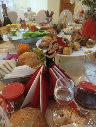 The photograph captures a festive indoor gathering, likely a banquet or celebration, set within a venue in Verkhnerusskoye, Russia. The focus is on a dining table laden with food and drink, arranged on a gold-colored tablecloth. Prominently featured is a decorative centerpiece depicting a flamenco dancer with a red fan, surrounded by red and white napkins.
To the left of the centerpiece, a basket holds flatbreads and buns, one of which is topped with sesame seeds. Next to this is a glass jar with a red lid, tied with red and white ribbon, containing colorful candies. In the foreground, two wine glasses are visible: one containing a clear liquid, likely water or white wine, and the other filled with a dark red liquid, presumably red wine.
The table extends into the background, showcasing a variety of dishes including what appears to be fried food, olives, sliced cucumbers, pickled vegetables, and shredded carrots. A bottle of champagne or sparkling wine stands on a tiered serving tray in the upper left corner. Several other bottles and containers are present, along with cutlery and other decorative elements, suggesting a well-prepared event. The lighting appears to be artificial, creating a warm and inviting atmosphere. The overall scene suggests a convivial occasion with ample food and a celebratory ambiance.