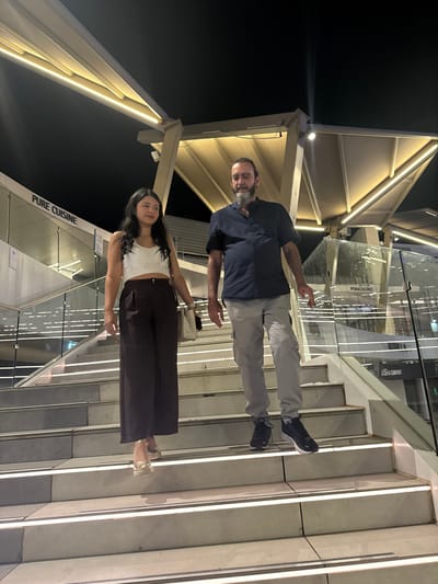 A young woman and a man are walking down a modern outdoor staircase at night. The woman, on the left, wears a white crop top and dark wide-leg pants. She carries a light-colored handbag. The man, on the right, is dressed in a dark short-sleeved collared shirt and light gray cargo pants, and wears dark sneakers. The staircase features illuminated strips along the edges of each step. In the background, there is a building with signage that includes "PURE CUISINE" and "POMODORO". The architecture of the surrounding structure is modern, with angular canopies and illuminated elements, suggesting a commercial or entertainment district. The location is identified as Alcorcón, Spain.