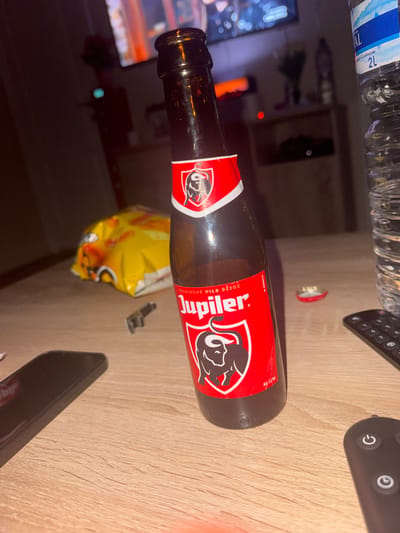 The image displays a dimly lit indoor scene on a light-colored wooden table surface, located in Temse, Belgium. Central to the foreground is an approximately half-full or recently consumed brown glass Jupiler beer bottle, standing upright. Its red label, featuring the brand name "Jupiler" and a bull's head logo, is clearly visible. To the right of the beer bottle, a clear plastic 2L water bottle with a "Val" brand label is present. Directly behind the Jupiler bottle lies a red metal bottle cap. To the left of the beer bottle, a crumpled yellow bag of potato chips is partially visible. Further to the left, a black smartphone is positioned, showing its rear side with a camera protrusion. A small, dark metallic object rests between the chip bag and the beer bottle. On the far right, a black remote control with white buttons is visible. The background is out of focus, showing a television screen displaying blurred lights and colors above a dark wooden cabinet or shelf unit. No individuals are present in the frame.
