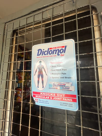 A sign for Diclo mol Pain Buster Gel is displayed behind a metal security grate in front of a store. The sign features the product name prominently, followed by "Knocks Out Muscular & Joint Pains." An anatomical diagram highlights areas of pain on a human figure. The sign lists indications for use: Joint Pain, Low Back Pain, Muscular Pain, and Sprains and Strains. An image of the product packaging is also visible on the sign, along with "Therapeutic Laboratories (N) Ltd." and an address in Maryland, Lagos. The store behind the grate appears to be stocked with various products on shelves. The scene is likely an outdoor view of a shop entrance, possibly in the city of Akowonjo, Nigeria, given the address details on the sign.