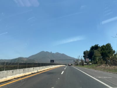 The image depicts a view from inside a vehicle traveling on a highway in San Cayetano, Mexico. The road is asphalt and marked with yellow lines on the left and white lane markers. A concrete barrier separates the highway from the shoulder on the left side of the frame. In the background is a mountain range under a clear blue sky. There is a small amount of vegetation on the right of the road. In the distance, another vehicle is visible on the same road.