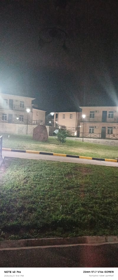 The image displays an outdoor scene at night, illuminated by artificial lighting. The foreground features a grassy area with a paved pathway bordered by yellow and black curbs. In the midground, there is a large, dark boulder and a small evergreen tree. Behind these elements, a low, weathered wall separates the grassy area from a slightly elevated section with two buildings.

The buildings appear to be residential or institutional, constructed with light-colored facades and multiple windows. Exterior lighting fixtures, resembling street lamps, are visible in front of the buildings. A dark, wrought-iron style railing runs along the elevated area. The background is dominated by a dark, nighttime sky. The text overlay at the bottom of the image indicates the photograph was taken with a "NOTE 40 Pro" device at "2026.06.23 11:41 PM" and displays camera settings "23mm f/1.7 1/14s ISO9878" along with the text "Nohijai tobei çumhuri".

This scene is likely located in Roghun, Tajikistan, as suggested by the text overlay. The overall impression is one of a quiet, possibly residential or recreational area at night. There are no people or animals visible in the frame, and no discernible actions or interactions are taking place. The focus is on the architectural elements, landscaping, and the atmospheric conditions of a night scene.