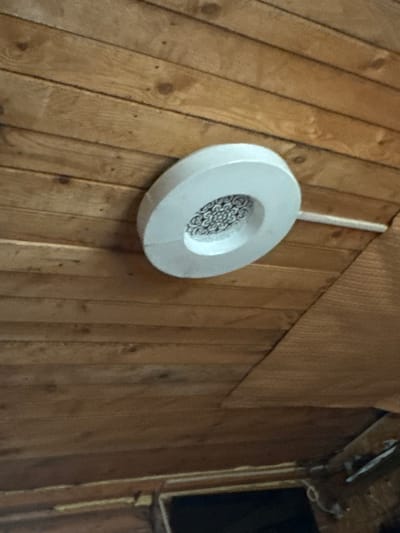 A white circular ceiling light fixture is mounted on a wooden plank ceiling. The light fixture has a decorative pattern in the center. A white conduit runs from the fixture across the ceiling. The ceiling is made of horizontal, light-brown wooden planks.